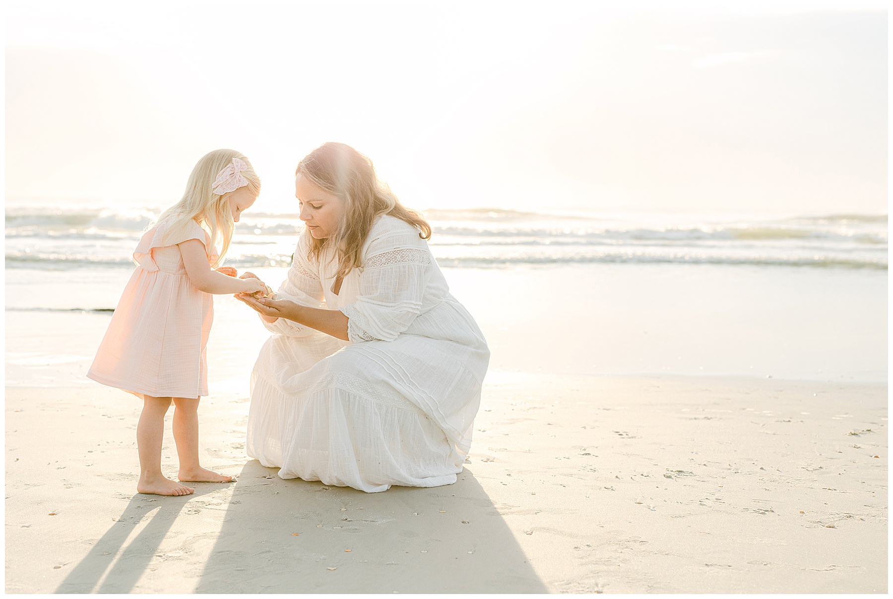 blond haired little girl kneeling with mom on the beach at sunrise in white dress light and airy light 