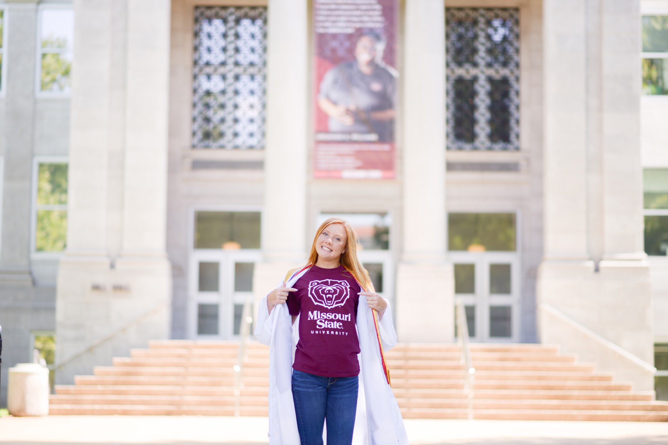 Senior girl in maroon tshirt and graduation robe standing in front of Missouri State University.