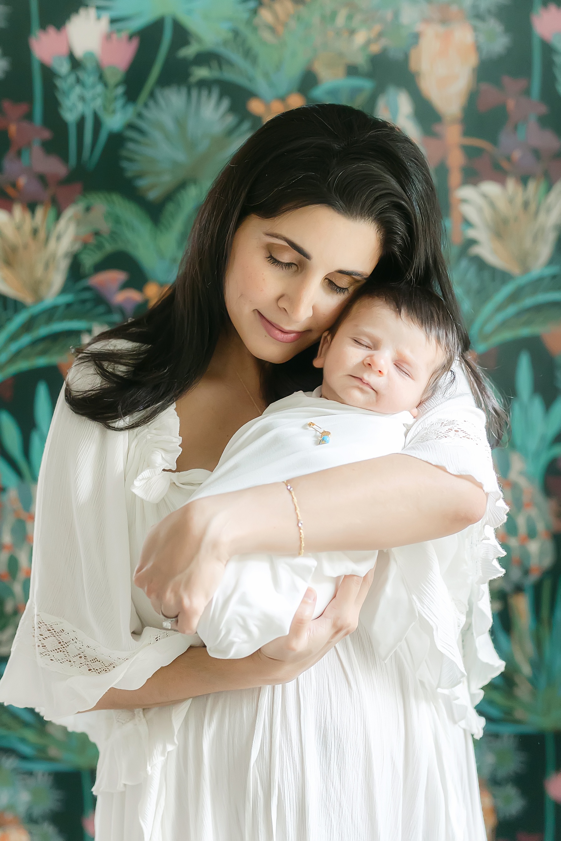 dark haired woman holding newborn baby boy wrapped in white swaddle against boho floral wallpaper