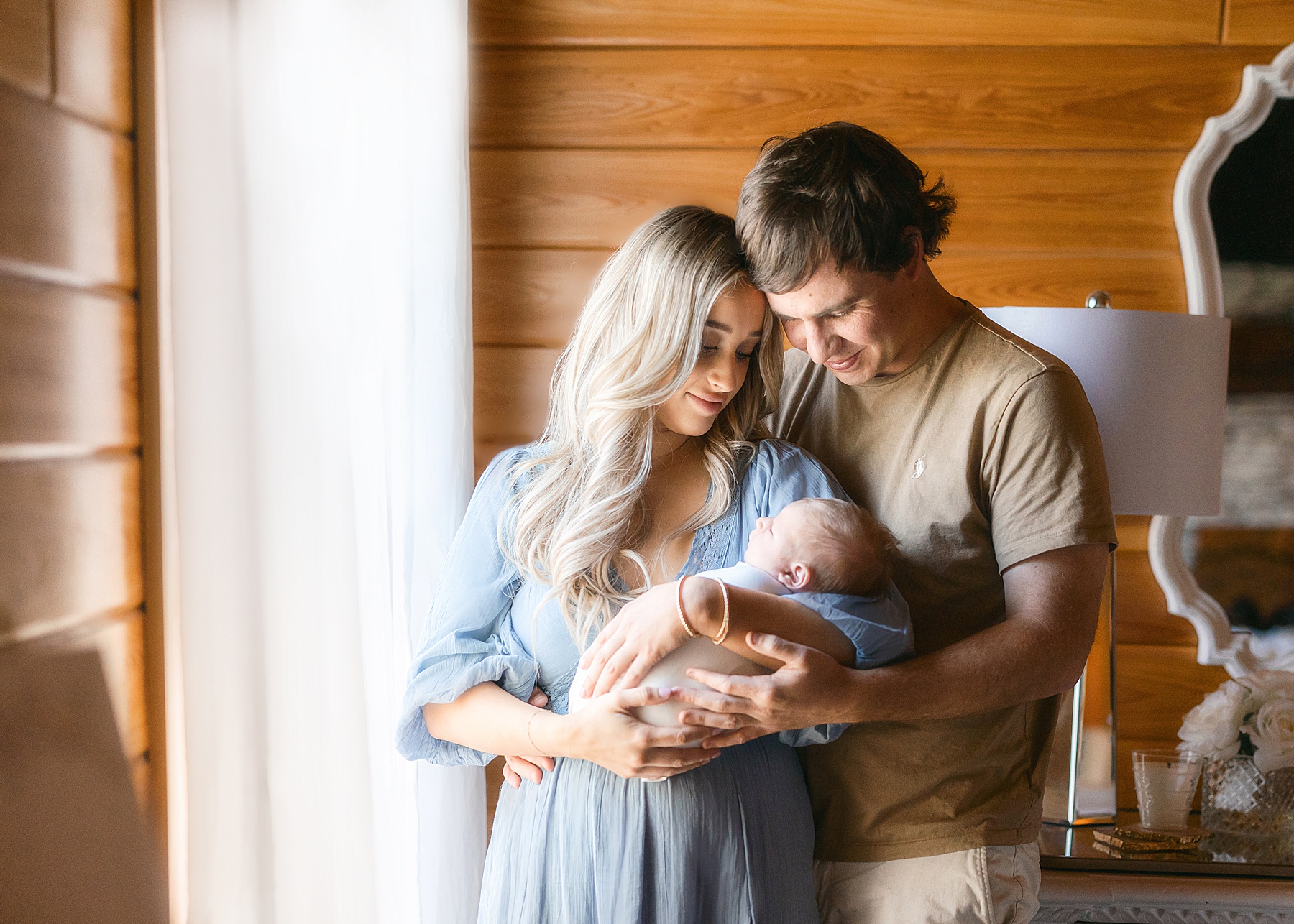 man and woman holding newborn baby boy in front of light and airy window with white sheers