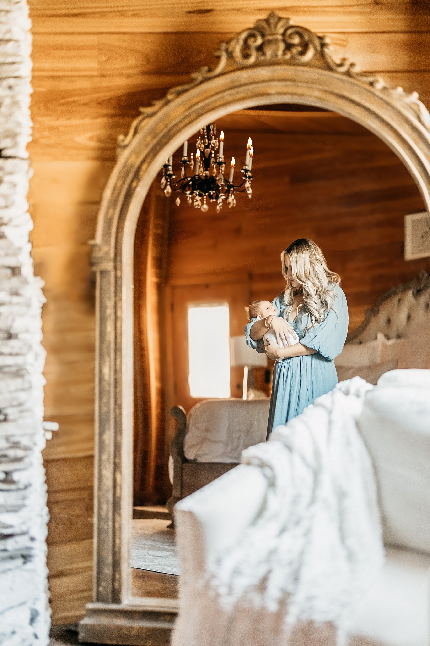 blond haired woman in maxi dress holding newborn baby boy in front of huge antique gold mirror