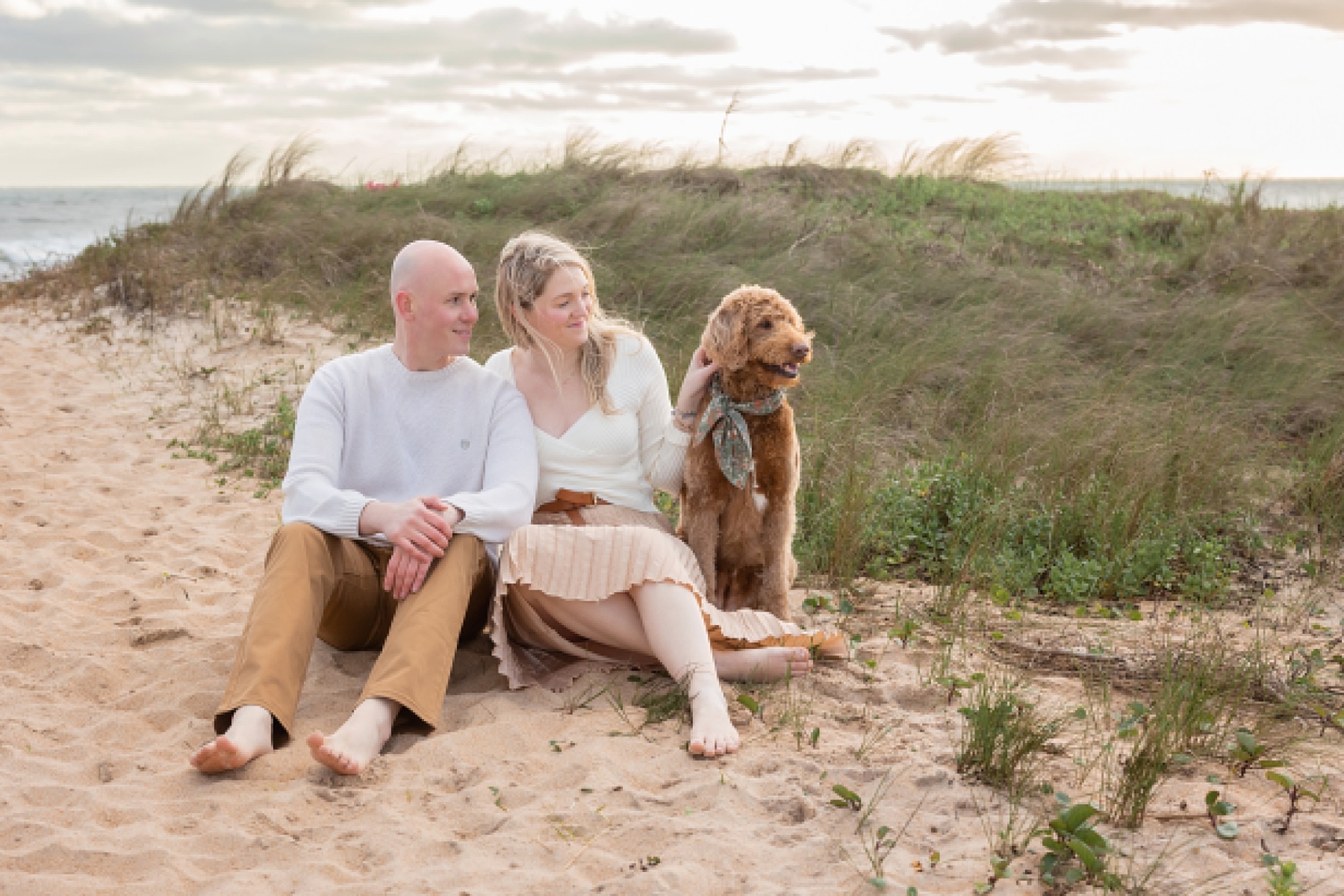 couple sitting on the beach in neutral colors with golden doodle