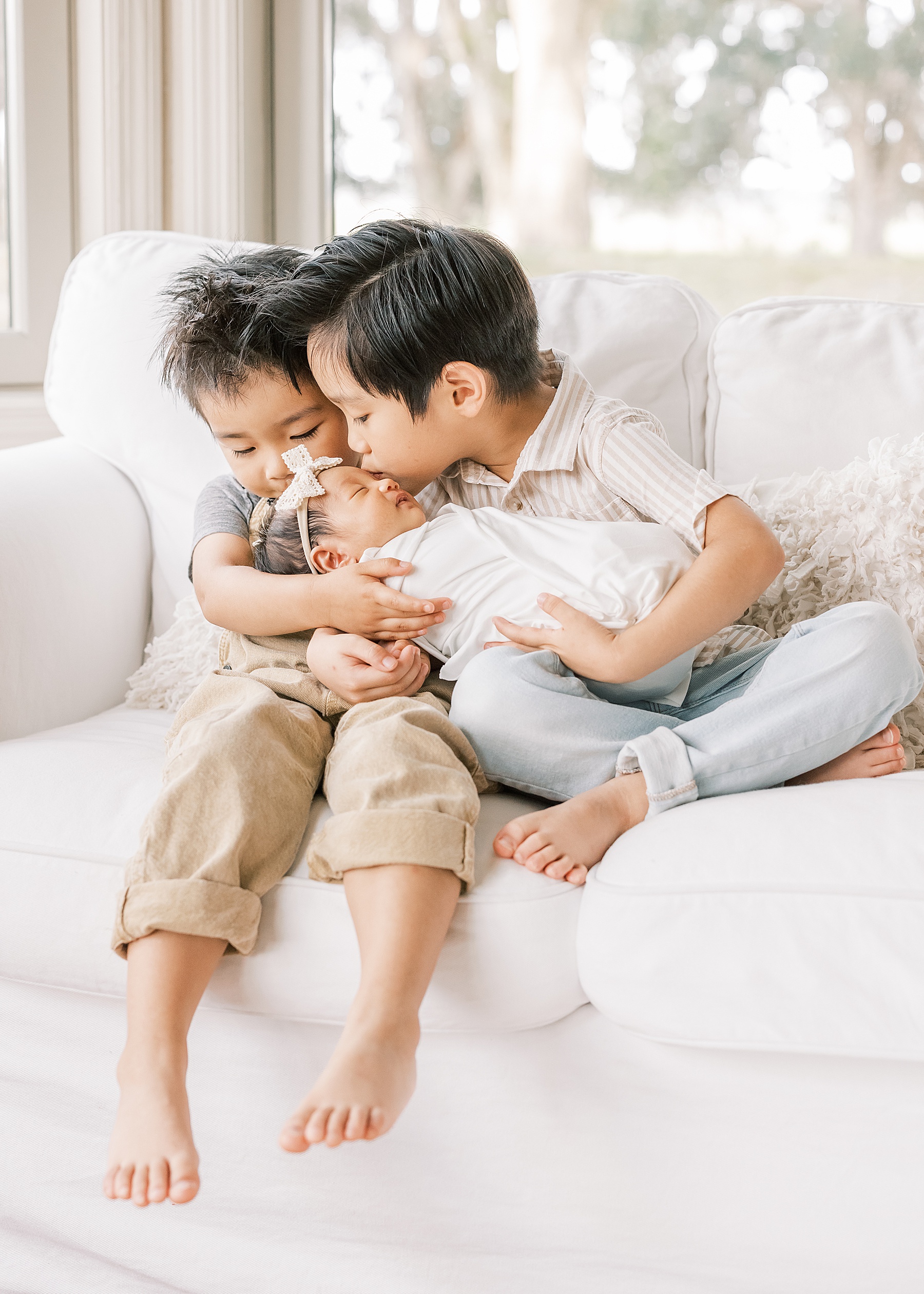 little boys dressed in neutral colors sitting on white couch kissing newborn baby sister