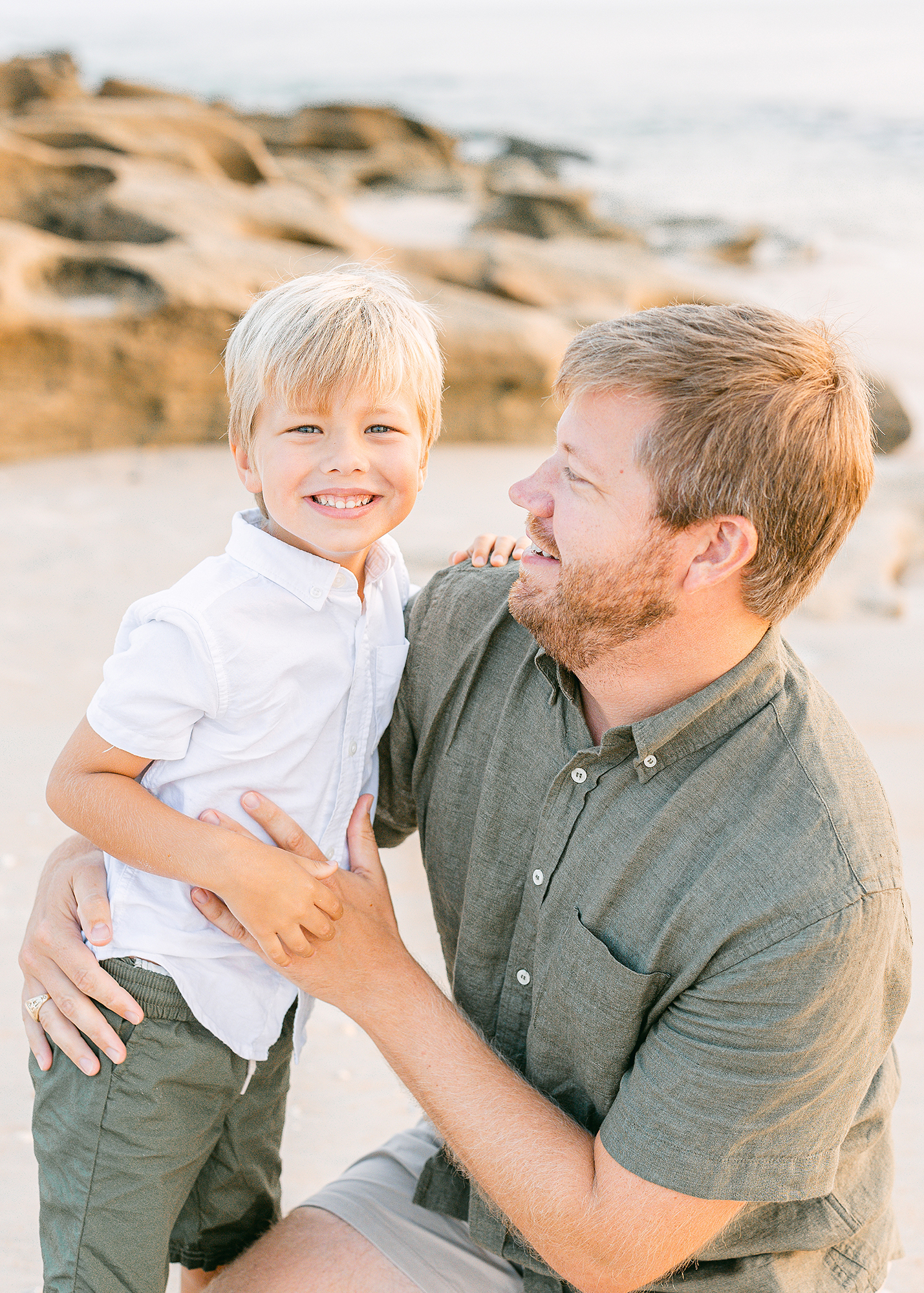 Man wearing neutral and sage green laughing with little boy on the beach at sunrise.