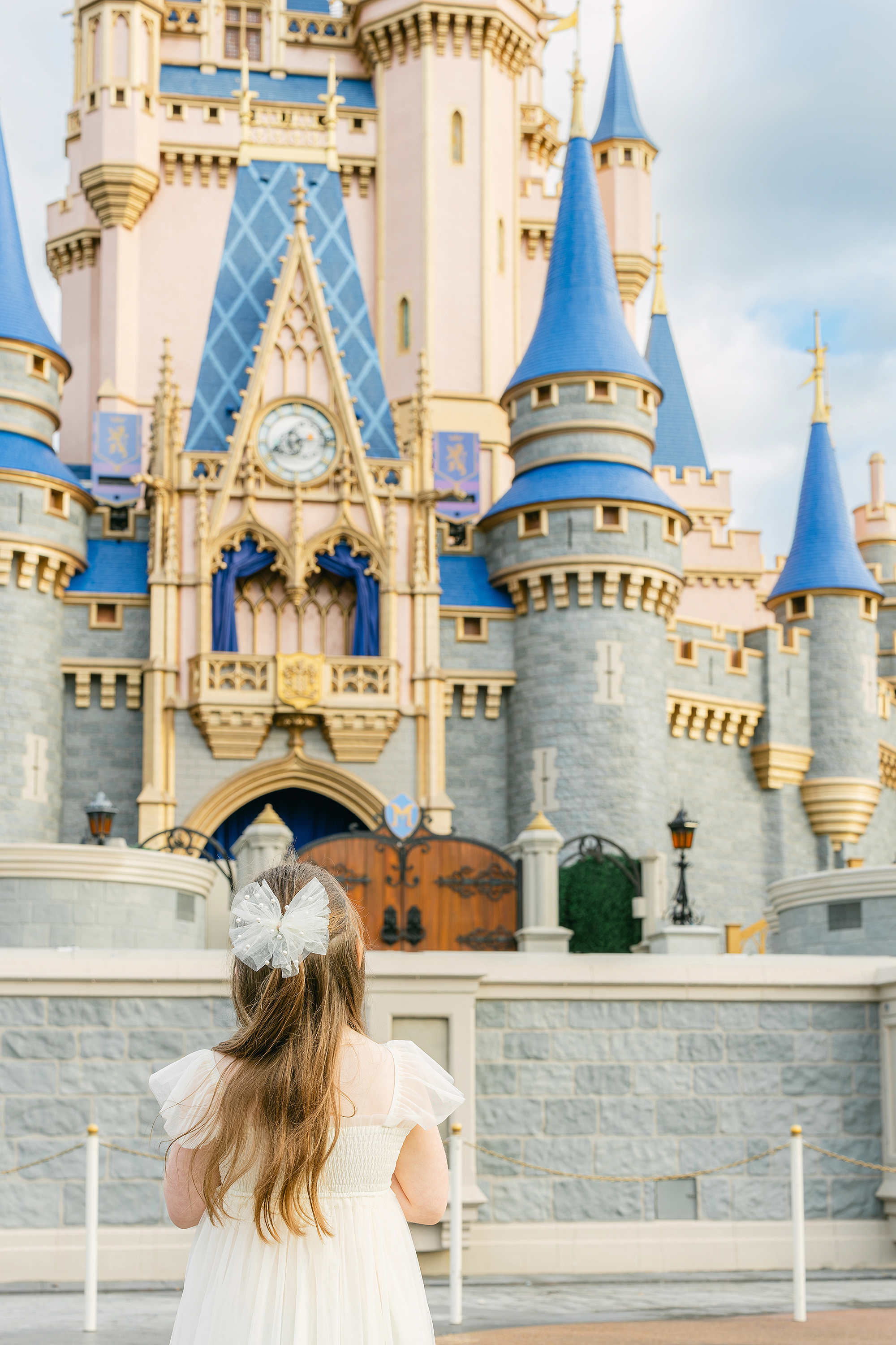A little girl in a cream dress with a cream bow looks out onto Cinderella's Castle.
