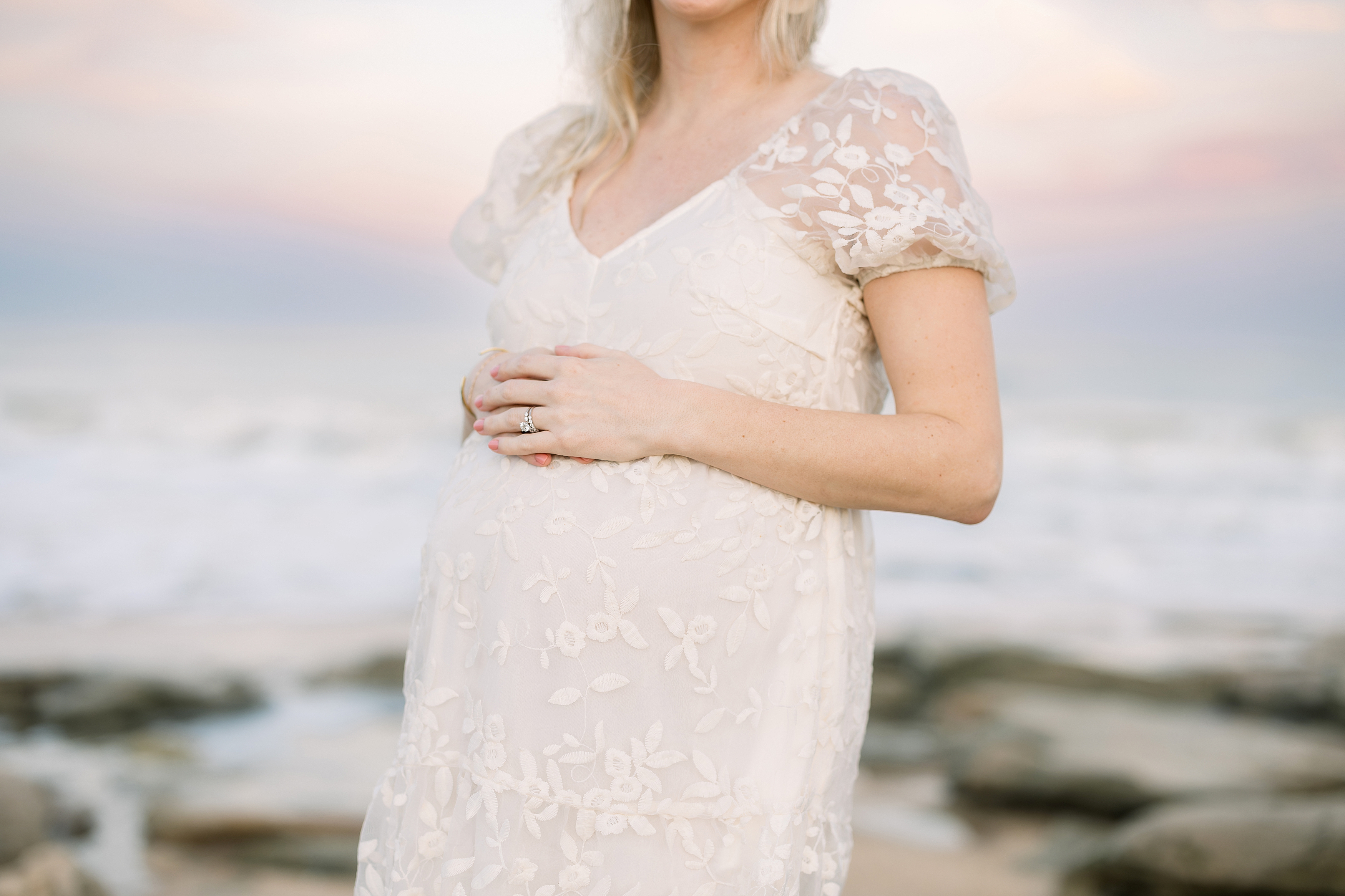 A pastel-hued sunset maternity portrait of a woman in a cream lace dress.