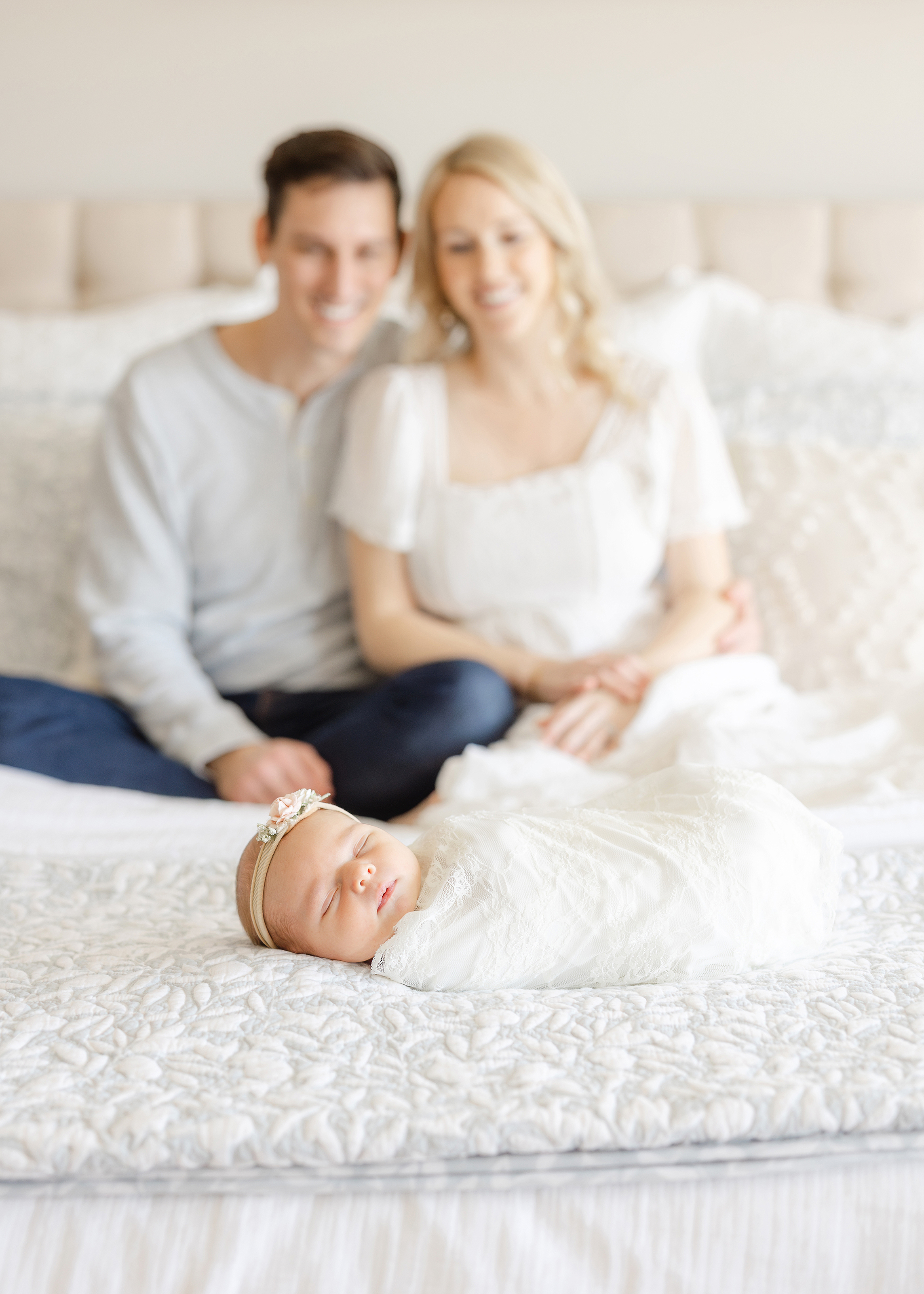 A newborn portrait of a couple sitting on their bed with their newborn baby girl.