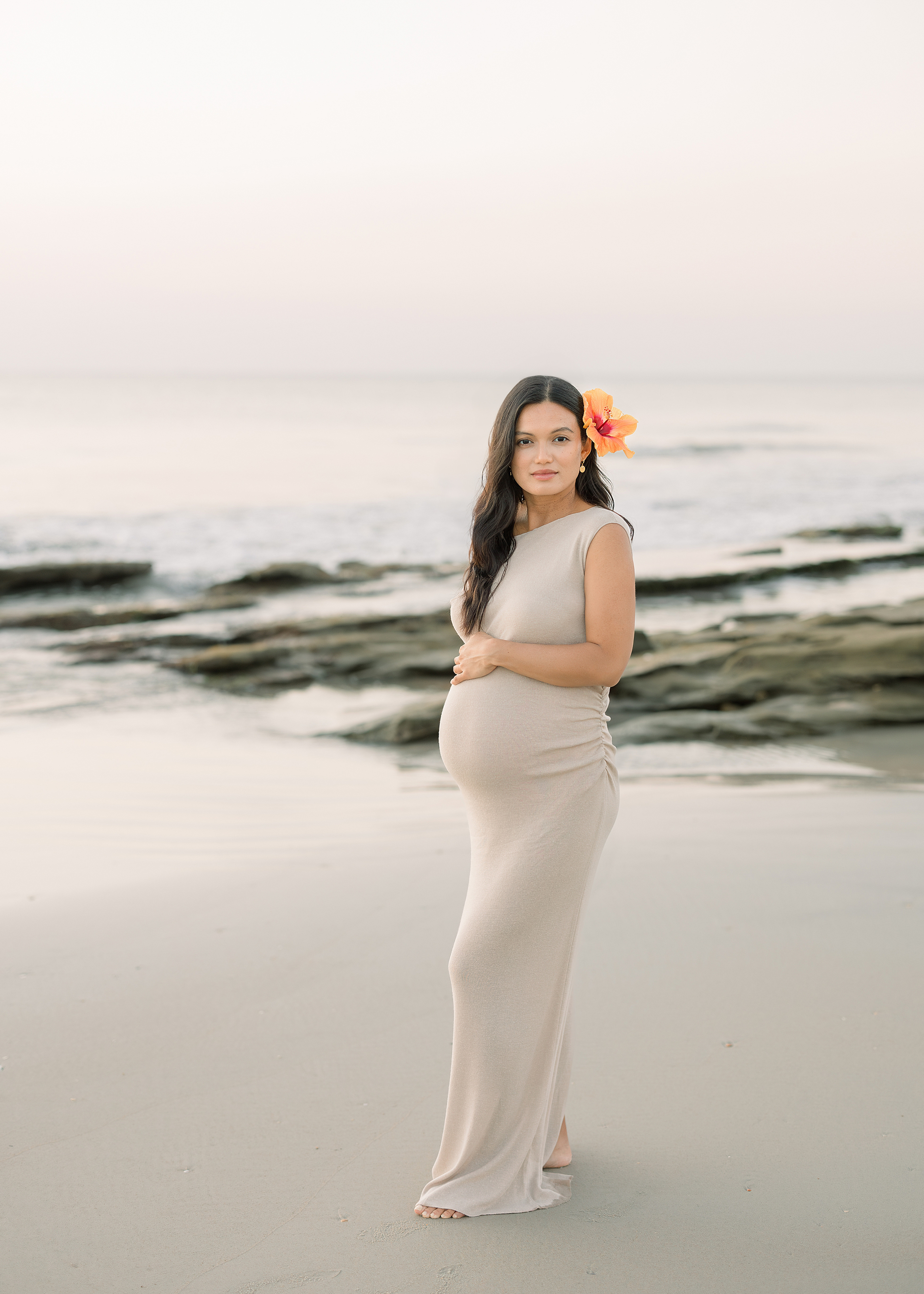 A sunrise pregnancy portrait of a woman in a neutral dress with a hibiscus flower in her hair.