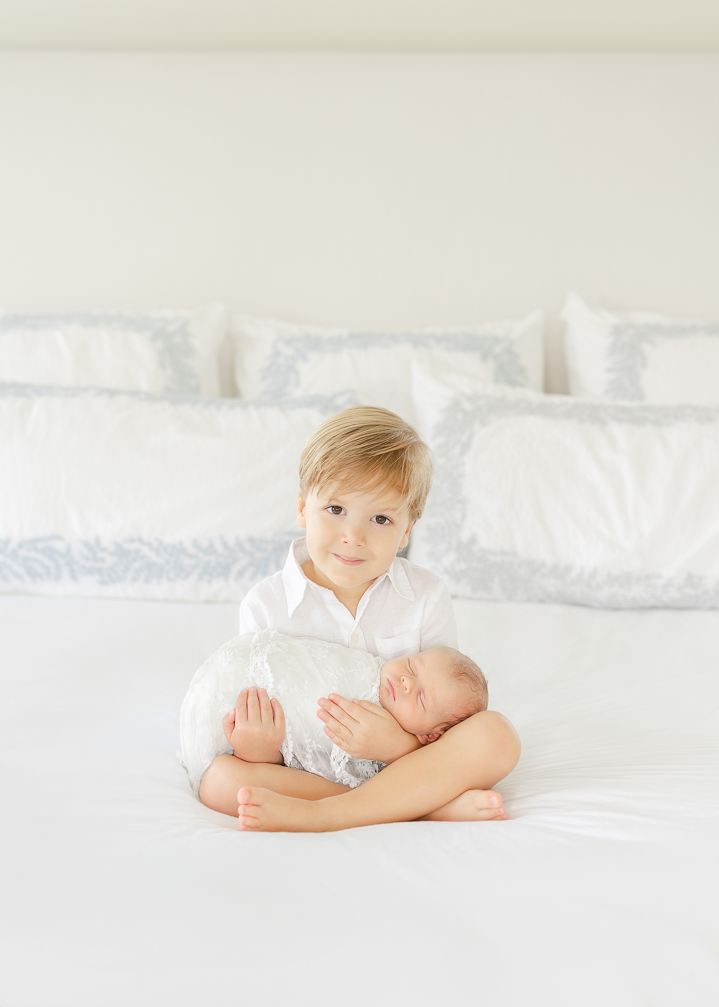 A toddler boy dressed in neutral colors holds his newborn baby sister on an all white bed during their in-home newborn session.