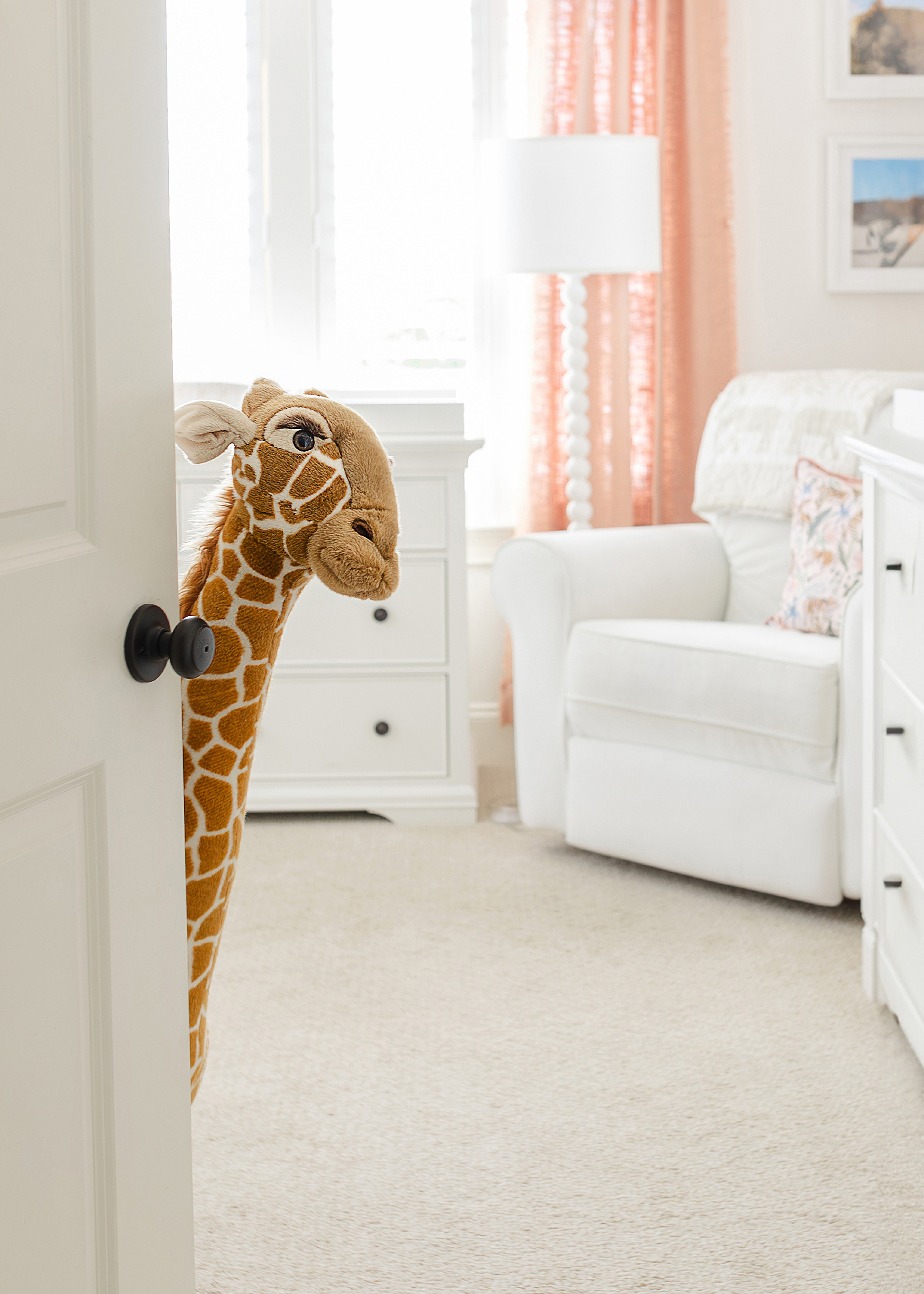 A pink and white newborn baby girl's nursery with a giraffe peeking out from the door as you enter the room.