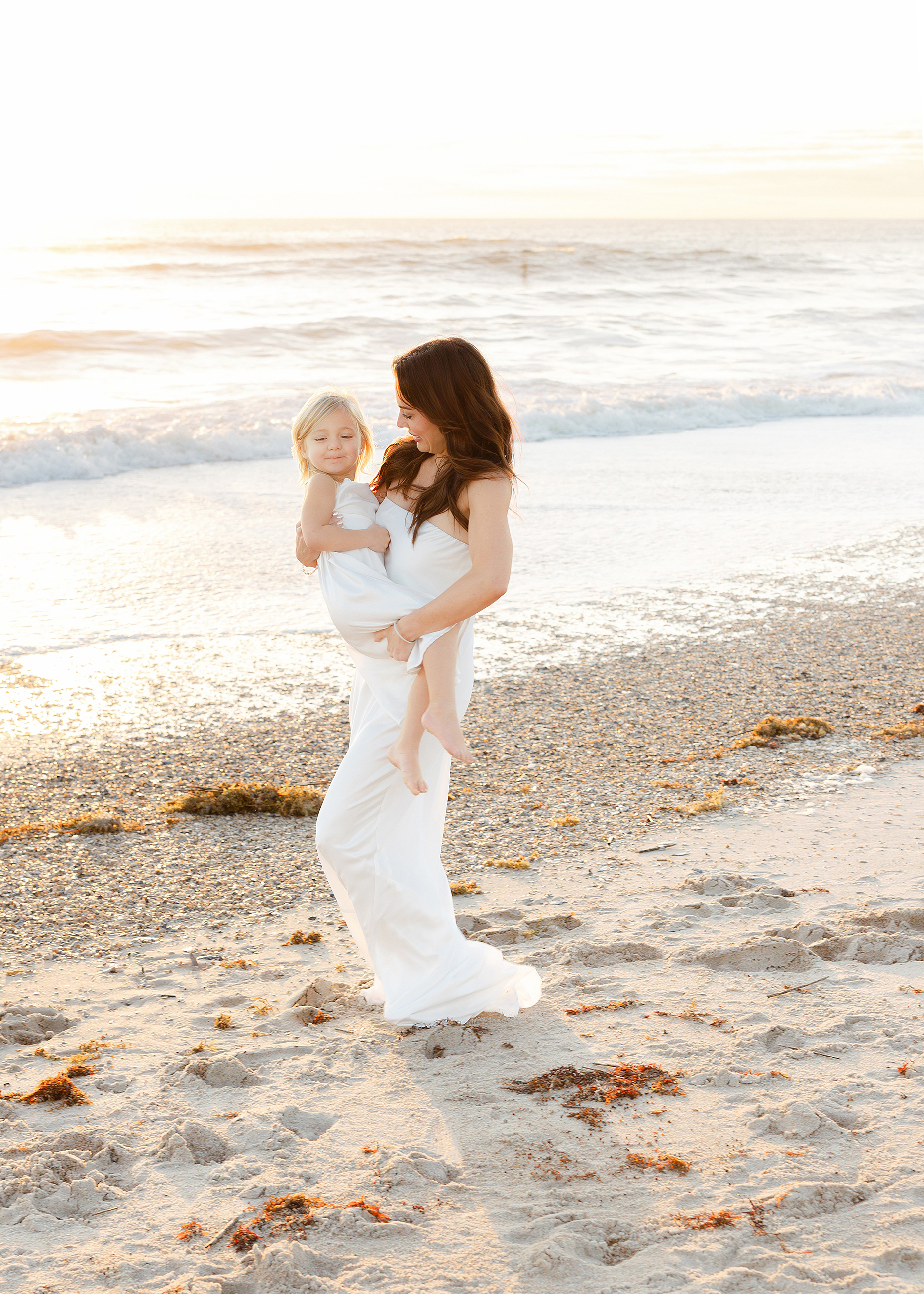 An airy sunrise portrait of a woman in a white dress holding a little girl on the beach.