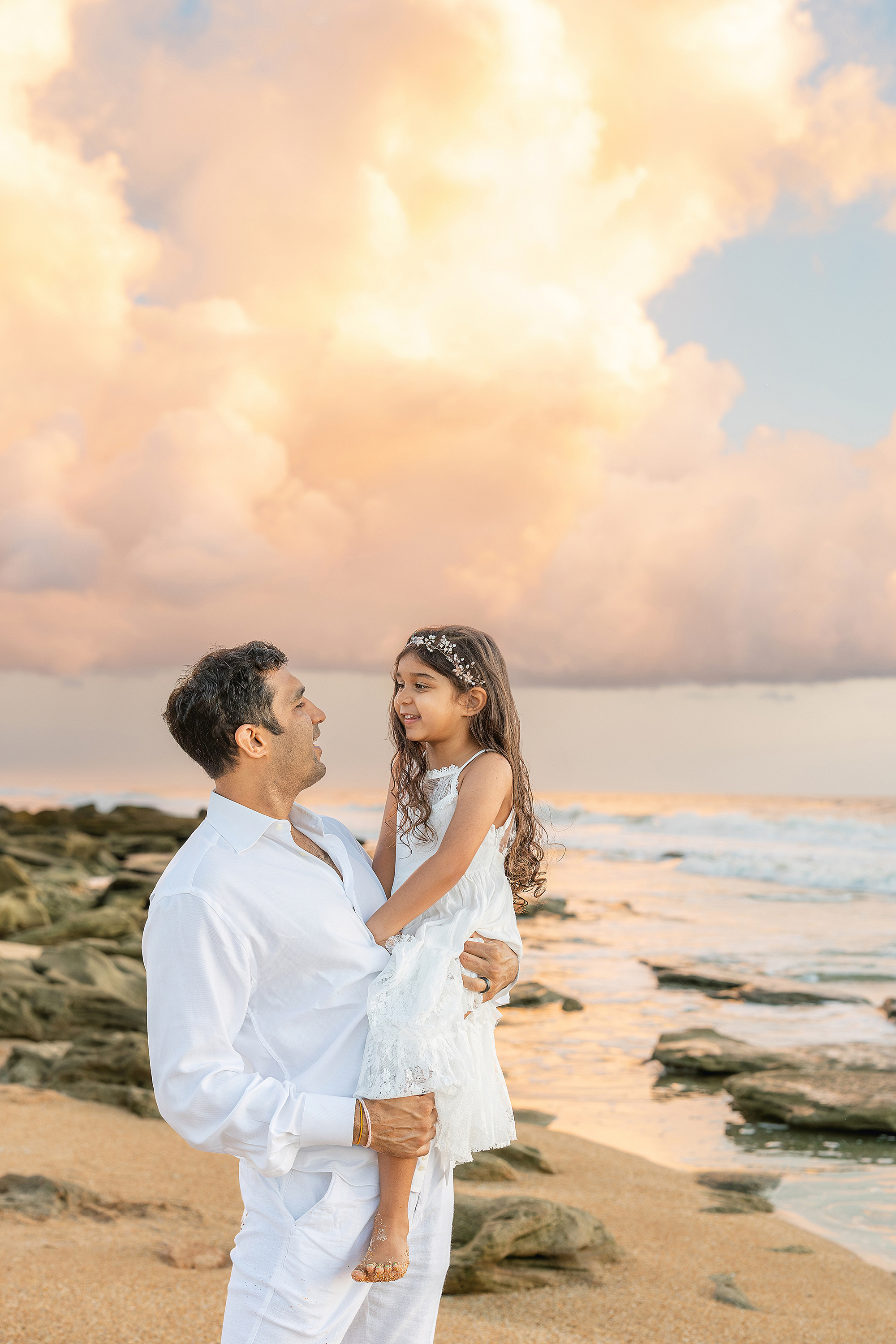 A man dressed in white holds his daughter on the beach during a colorful, pastel sunrise.