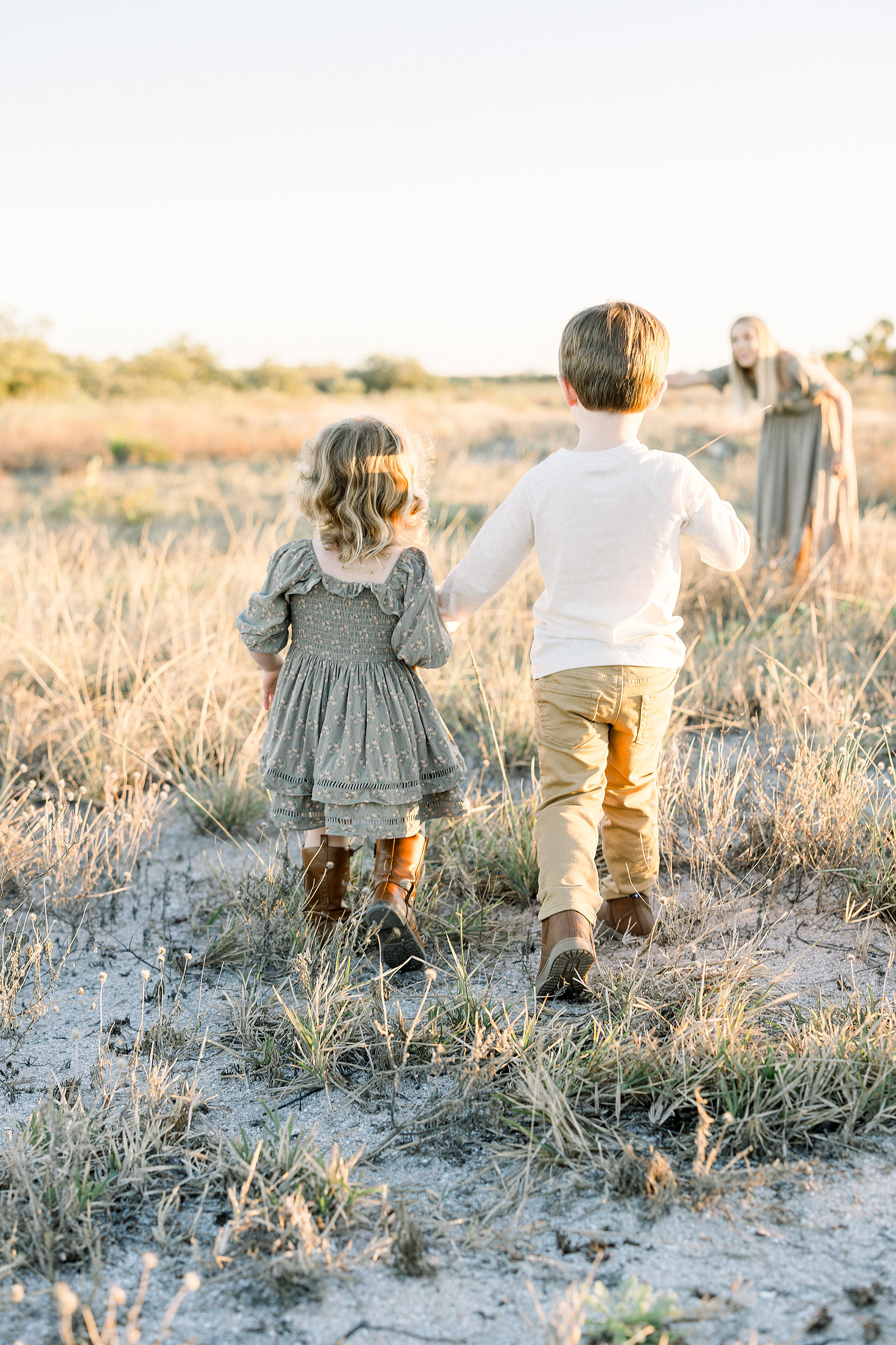 Two children hold hands and walk towards their mother in a backlit late afternoon sunshine near the beach.