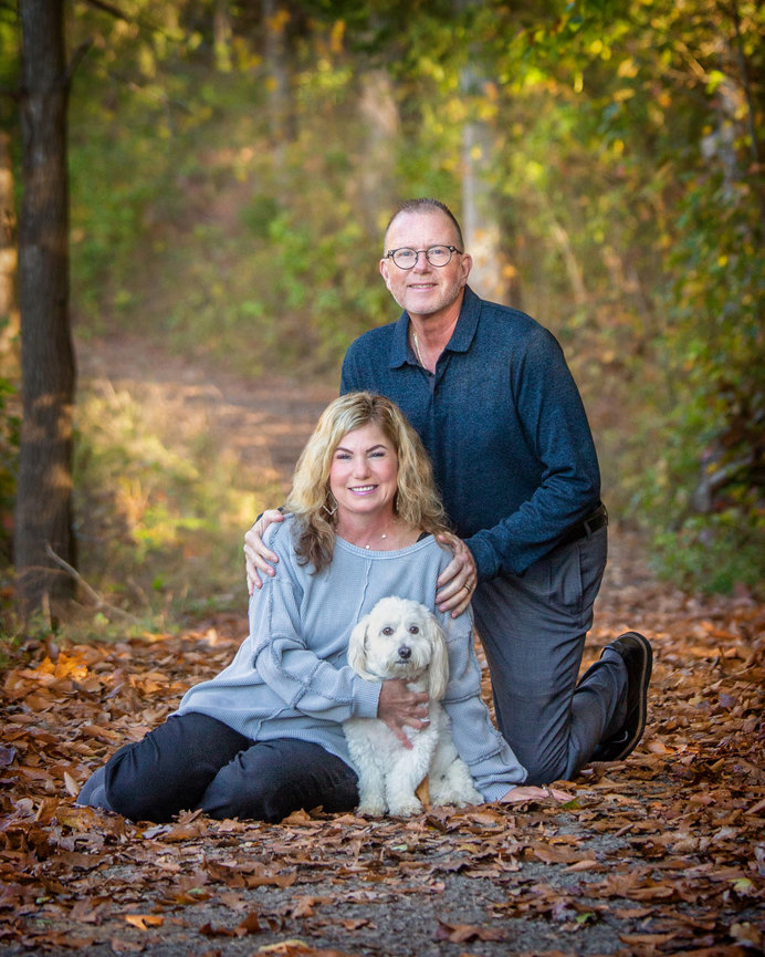 Couple kneeling on a forest path with a small white dog, surrounded by autumn leaves.