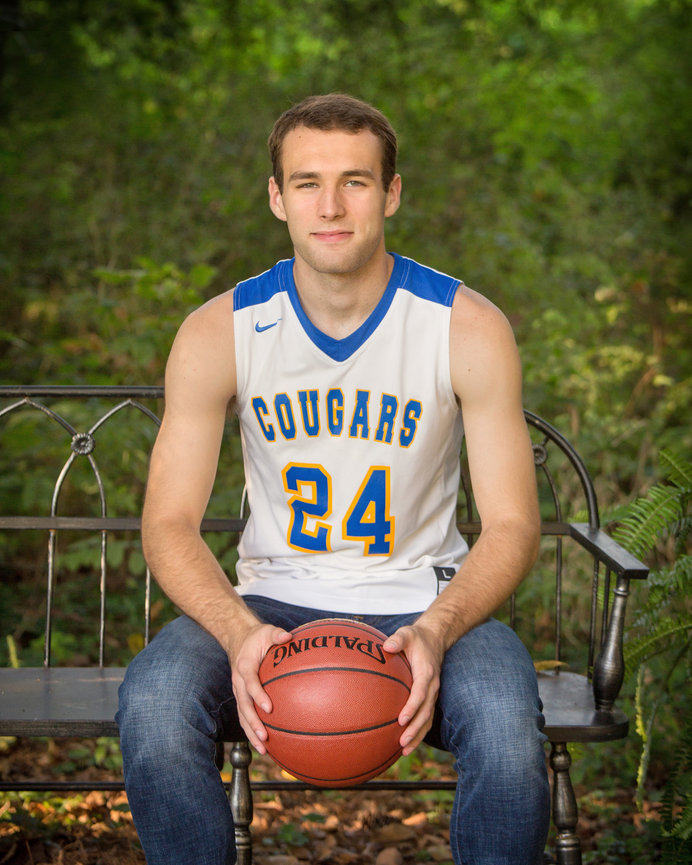 Basketball player in Cougars jersey holding a ball, sitting on a bench in a lush, green outdoor setting.