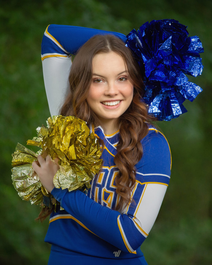 Cheerleader in blue and yellow uniform holds matching pom-poms with a green foliage background.