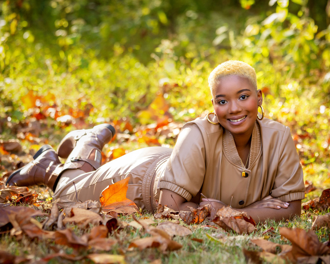Person with short hair lying on autumn leaves in a sunlit park, wearing a tan outfit and boots.
