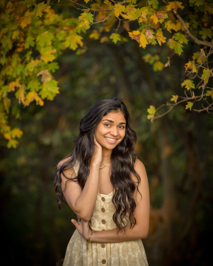 Woman in a beige dress smiles, standing amidst green and yellow leaves.