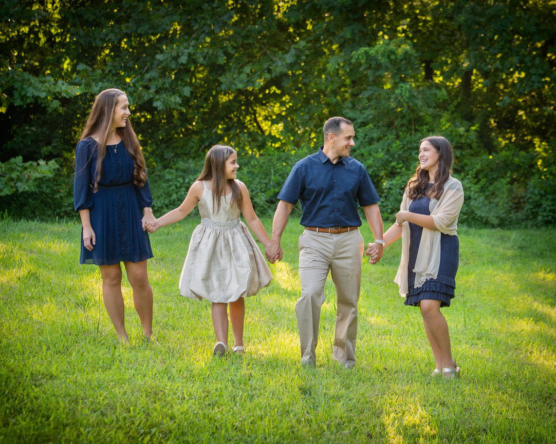 Family of four walking on grass, holding hands, smiling, with trees in the background.