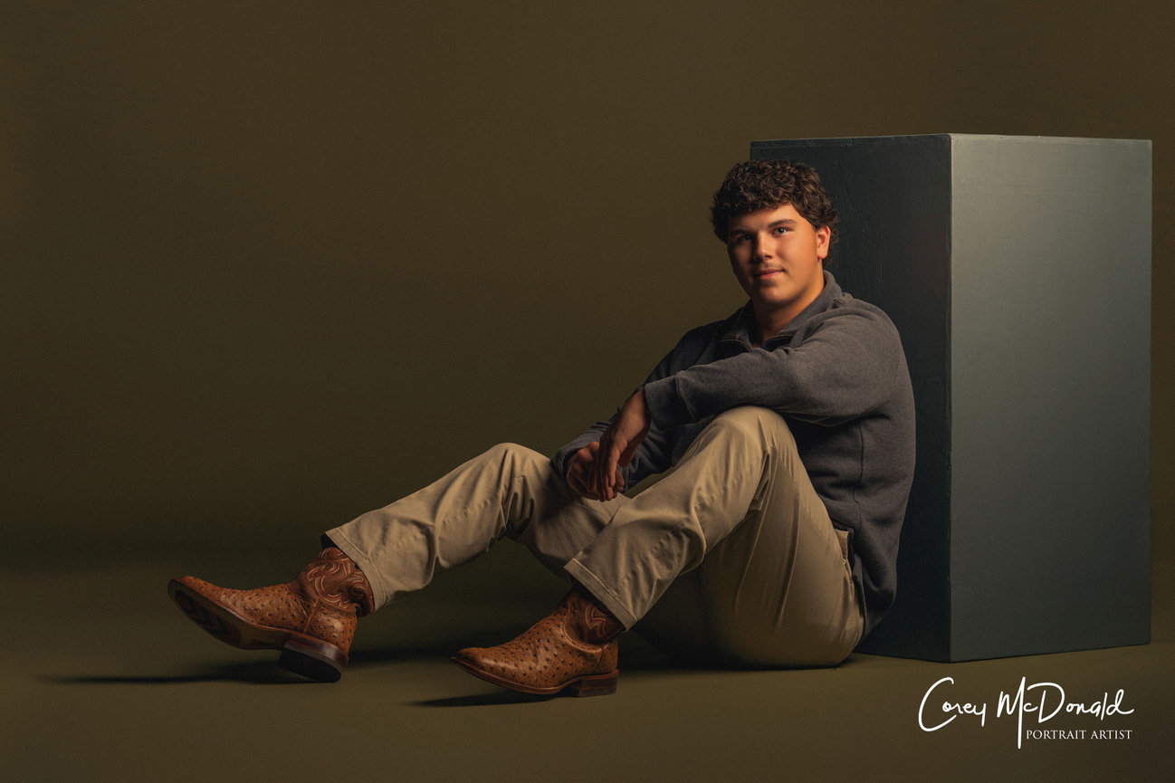 Young person sitting on the floor, leaning against a box, wearing a sweater, khaki pants, and cowboy boots.