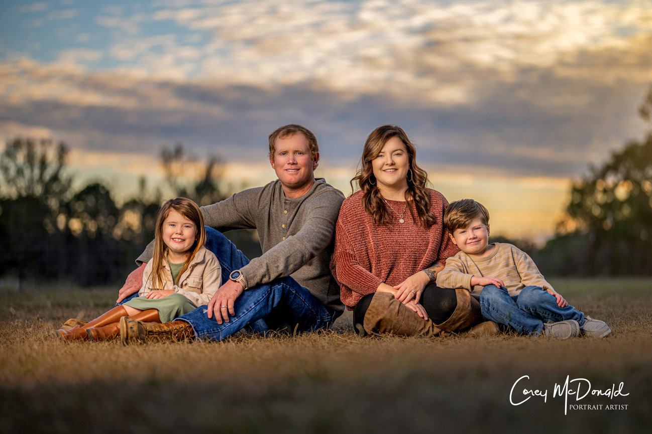 Family sitting on grass in a field at sunset, with a cloudy sky backdrop. Parents and two children smiling.