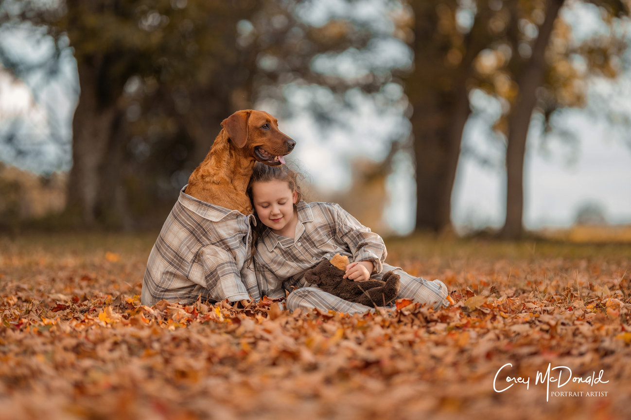 Girl and dog in matching outfits sitting on autumn leaves with trees in the background.