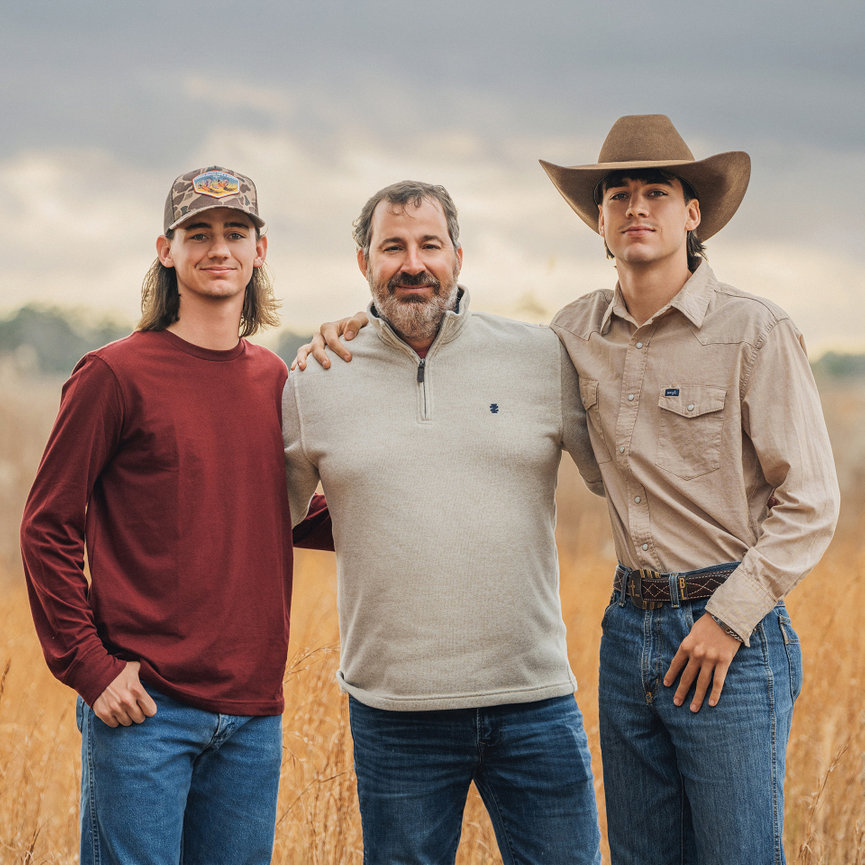 A man in a sweater stands between two young men in casual attire with a field in the background.