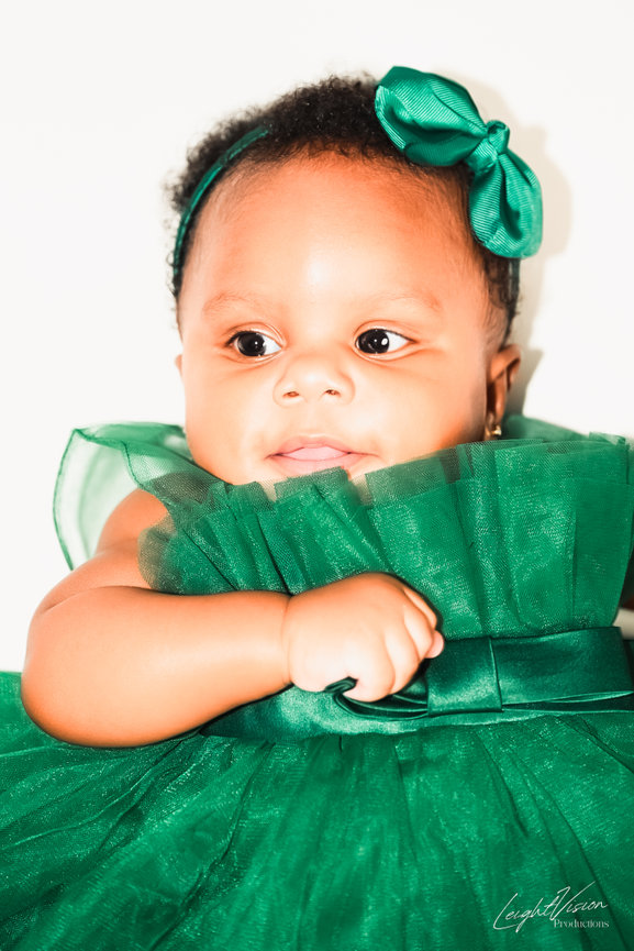 A baby girl wearing a green dress adorned with a bow, smiling joyfully in a bright, cheerful setting.