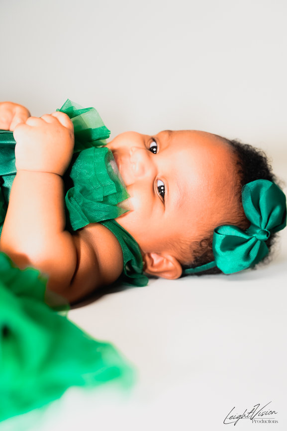 A baby girl in a green dress is positioned on her stomach, radiating cuteness and joy in her playful pose.