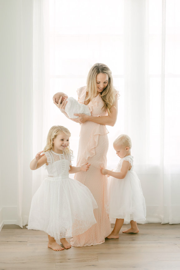 A woman in a pale pink dress holds her newborn baby girl while her two other girls dance around her.
