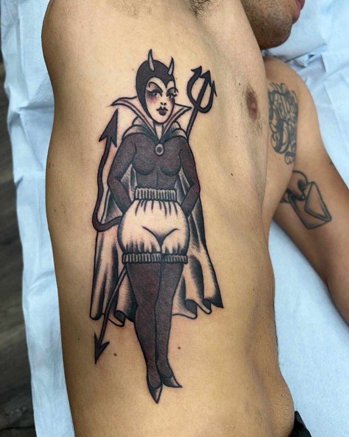 Tattoo on a person's side depicting a she-devil character with horns, a pointed tail, holding a trident, and wearing a cape and form-fitting outfit with stockings. The tattoo is done in a traditional black & gray style and was made by Beam.