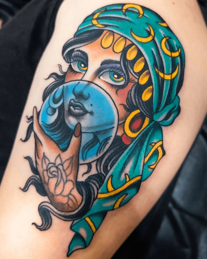Colorful tattoo of a fortune teller wearing a blue headscarf with gold coins holding a crystal ball.