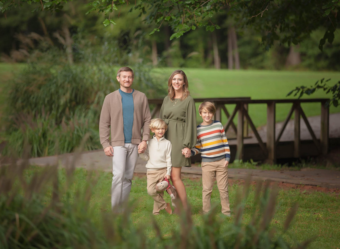 Family of four posing outdoors in front of a small wooden bridge, captured in soft natural light with a timeless style.