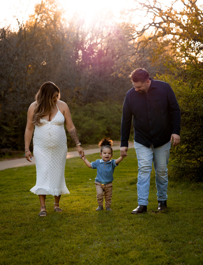 A family walks hand in hand on grass under a sunset sky, with trees in the background.