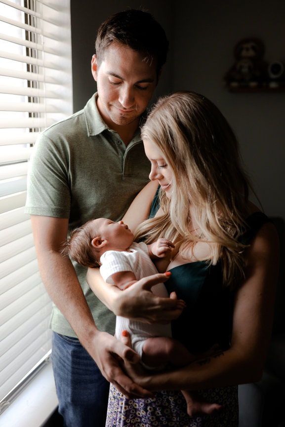 A couple lovingly holds their sleeping newborn near a window, bathed in soft natural light.