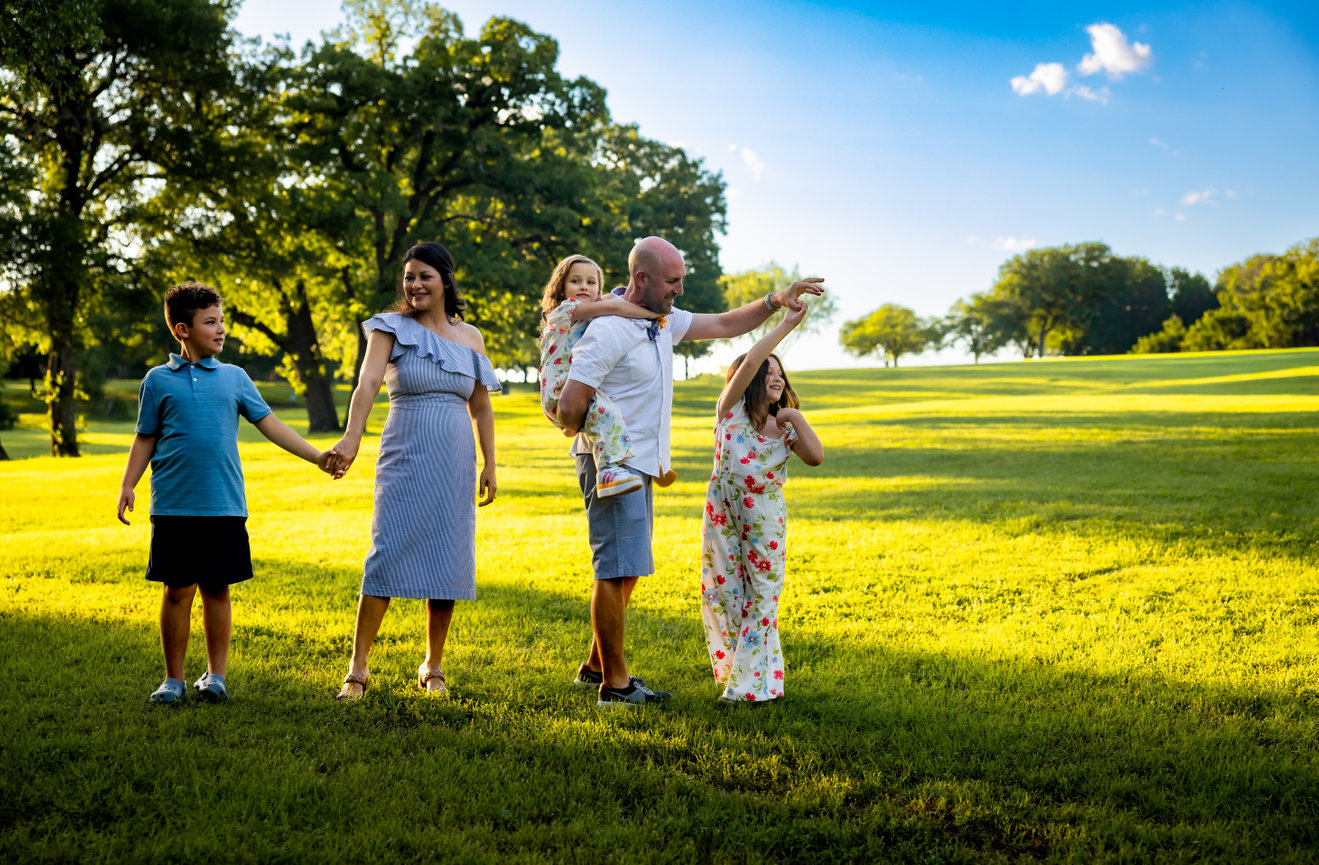 Family of five playing on a sunny park lawn with trees and blue sky