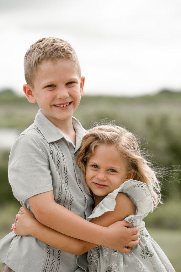 Two smiling children embrace outdoors, surrounded by greenery, with a cloudy sky in the background. Iowa City, terry True Blood Recreation Area, Familly photography