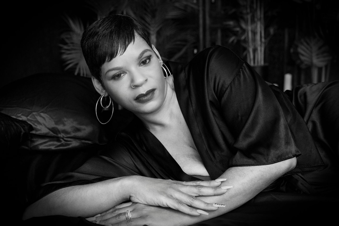 Woman in a black satin robe lounges gracefully, with a thoughtful expression and hoop earrings in a softly lit setting.