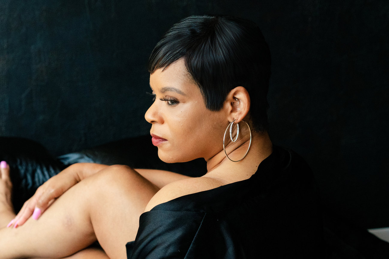 Woman with short hair and hoop earrings seated on a dark background, looking over her shoulder.