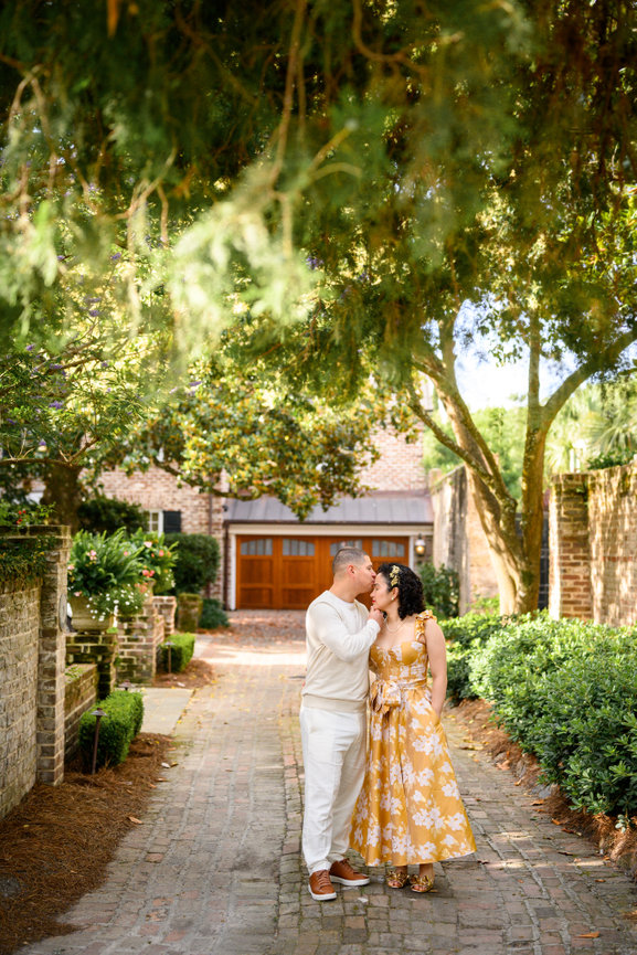 A couple standing on a cobblestone path under trees, smiling at each other; yellow dress and light outfit.