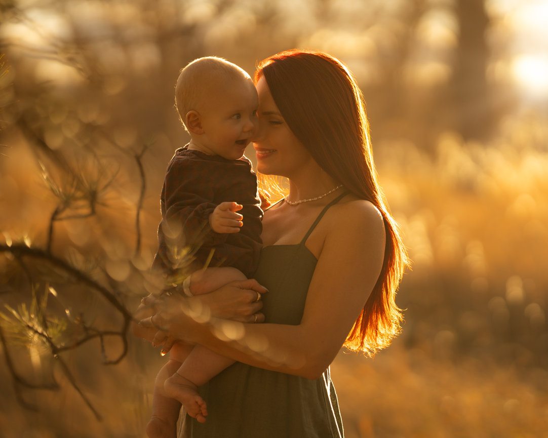 A woman with long red hair holds a baby outside in a sunny, golden field, in Lakewood, Colorado.