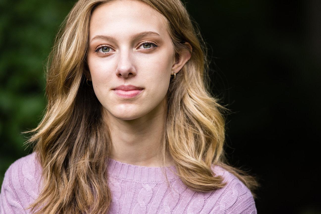 High school senior girl with long blond hair and a pink sweater. She is looking at the camera with a small smile.