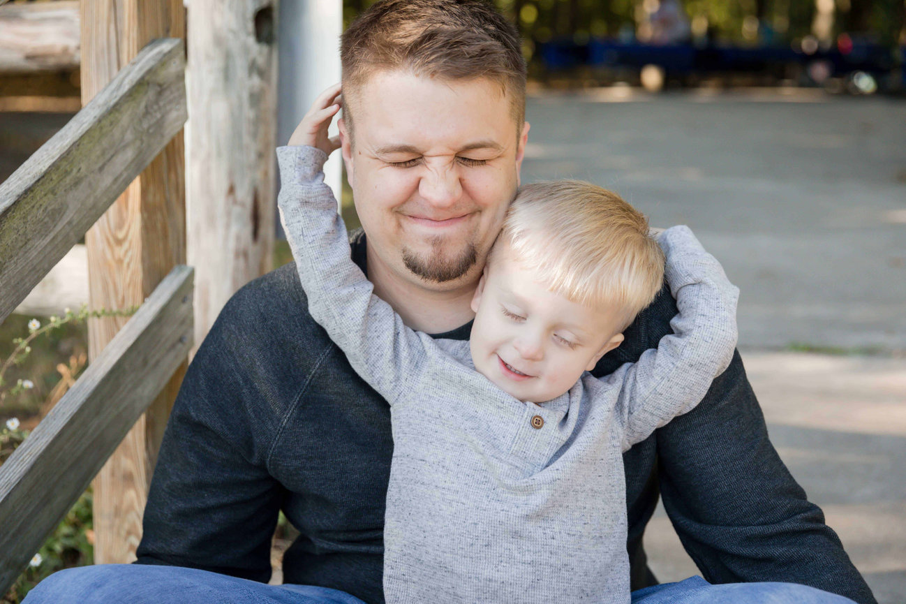 Father hugging smiling young child outdoors on a wooden walkway during family photo session in Blue Springs photographed by Case Studios Photography