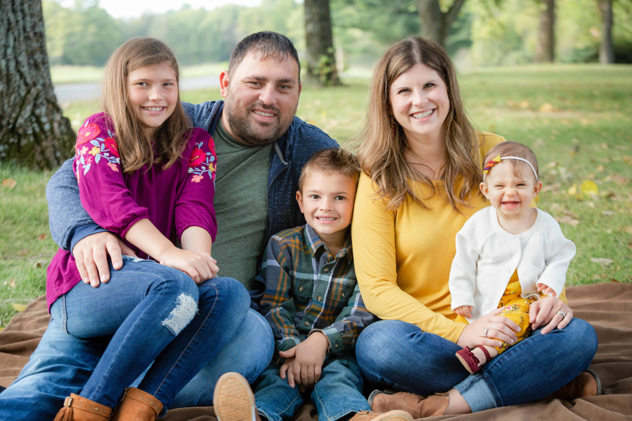 Smiling family of five sitting on a blanket in a park with trees in the background in a Blue Springs family photo session photographed by Case Studios Photography