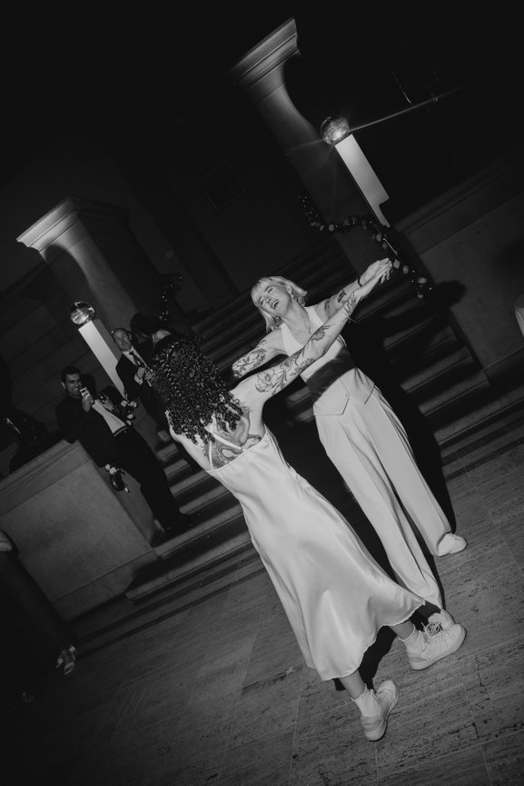 Black and white flash photography of first dance at Manchester Art Gallery, capturing reaction of a couple of guests stood on the stone stairs behind the two brides.