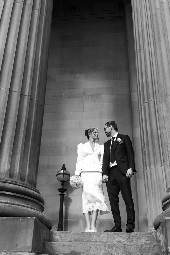 Editorial elopement photography of a bride in a vintage white suit and birdcage veil staring at her groom, framed by the grand stone columns of St George's Hall, Liverpool.