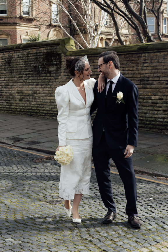 Editorial wedding portrait of a bride in a vintage jacket and skirt holding white roses, walking with her groom on a Liverpool cobblestone street in natural light.