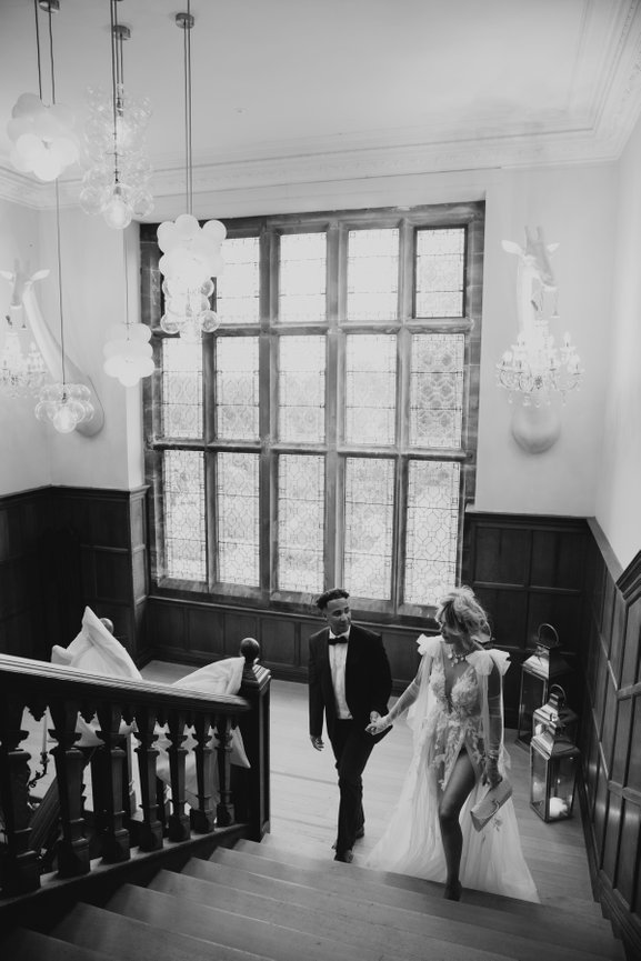 Editorial wedding photography: Bride, Tara Maynard, dress in long wedding dress walks up the grand stair case of The Salt House, with groom, Nicky Maynard, in black tie for their wedding anniversary at Standon Hall.
