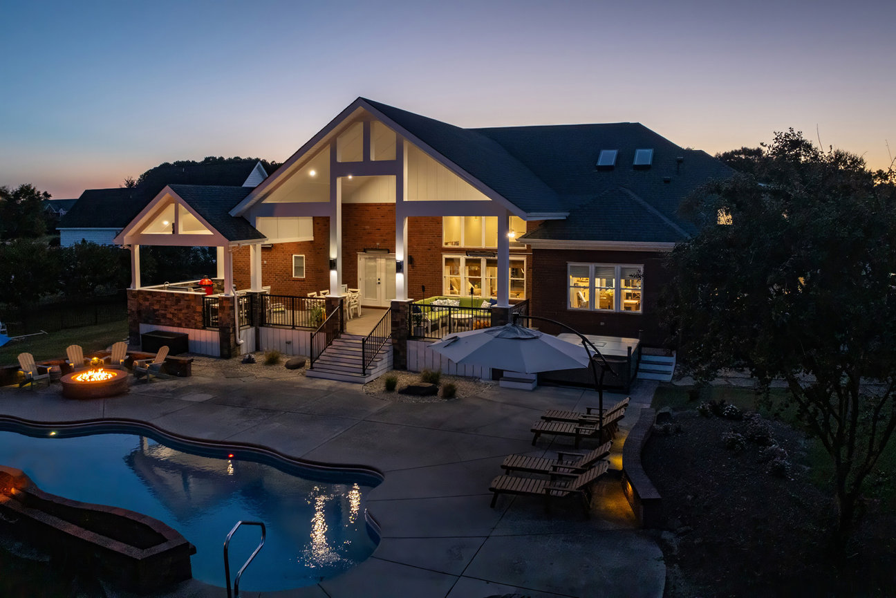 Spacious house at dusk with a lit patio, pool, lounge chairs, and a fire pit in the backyard.