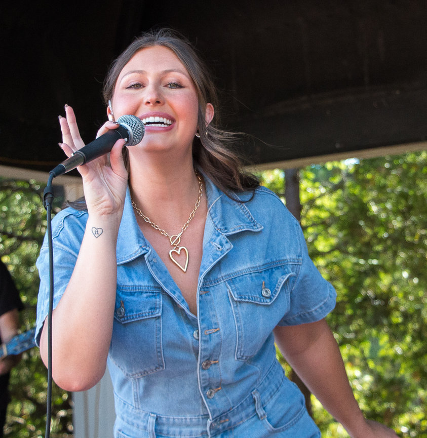 A woman in a denim outfit sings into a microphone outdoors, with trees in the background.